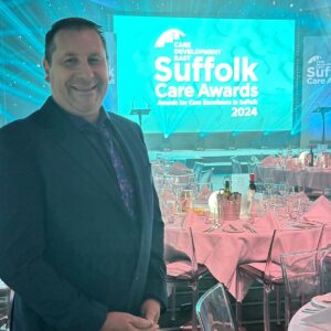 Alan, a support worker for Thera East, wins 'Runner Up' at the Suffolk Care Awards 2024. Alan is in the foreground of the photo with the Suffolk Care Awards Presentation in the background.