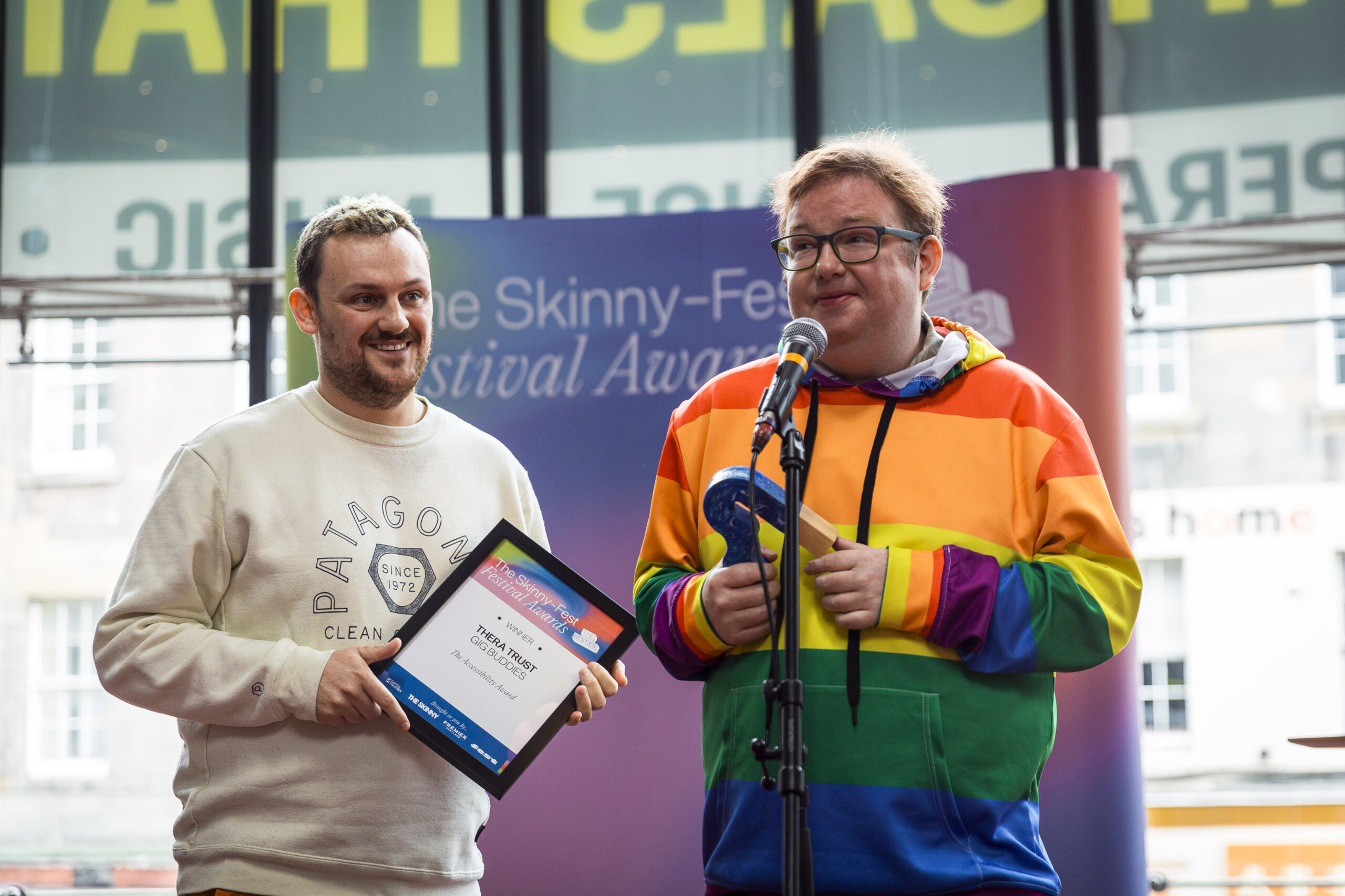 Sam is smiling onstage holding a certificate with Joseph who is speaking on the microphone at the 2024 Skinny-Fest Festival Awards. 